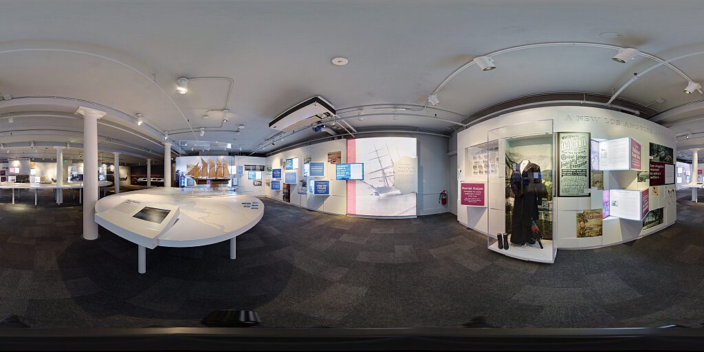 Panoramic view of Improbable Gateway Exhibit showing large wall photos, pop out text panels and in-depth descriptions of the transportation story of the Los Angeles Harbor. Artifacts include a tall ship replica and a model steam train.