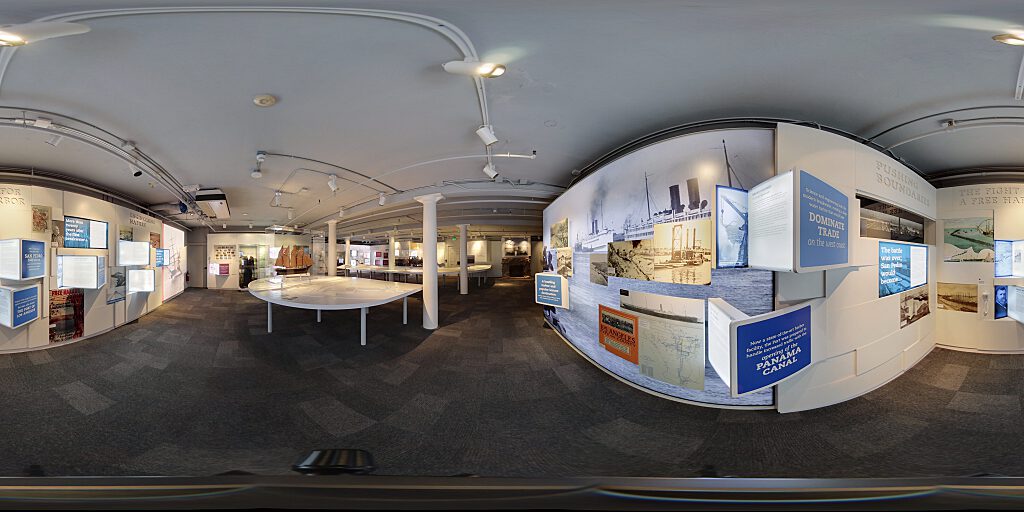 Panoramic view of Improbable Gateway Exhibit showing large wall photos, pop out text panels and in-depth descriptions of the transportation story of the Los Angeles Harbor. Artifacts include a tall ship replica and a model steam train.