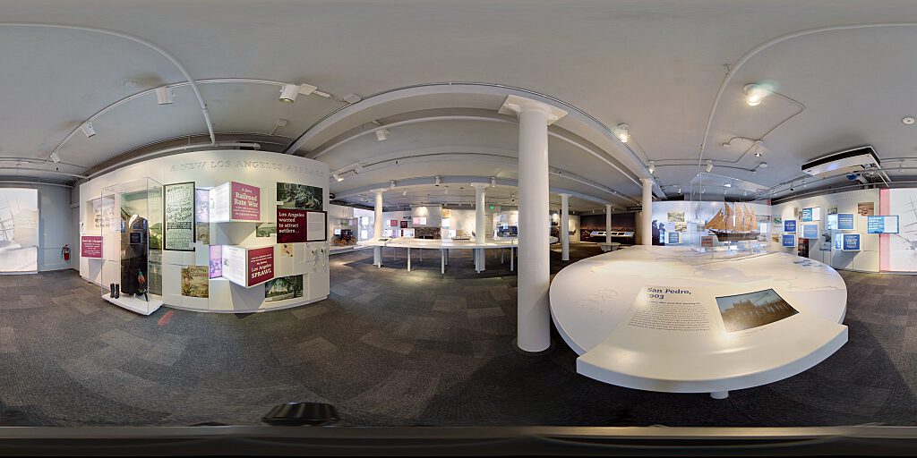 Panoramic view of Improbable Gateway Exhibit showing large wall photos, pop out text panels and in-depth descriptions of the transportation story of the Los Angeles Harbor. Artifacts include a tall ship replica and a model steam train.