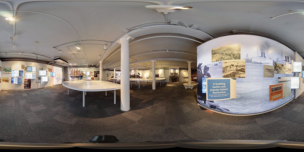 Panoramic view of Improbable Gateway Exhibit showing large wall photos, pop out text panels and in-depth descriptions of the transportation story of the Los Angeles Harbor. Artifacts include a tall ship replica and a model steam train.
