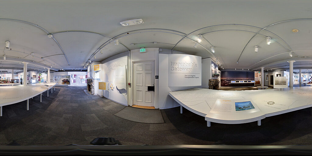 Panoramic view of Improbable Gateway Exhibit showing large wall photos, pop out text panels and in-depth descriptions of the transportation story of the Los Angeles Harbor. Artifacts include a tall ship replica and a model steam train.
