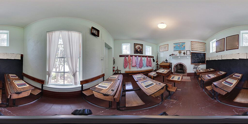 Panoramic view of Schoolhouse interior with desks with books, pinafores, and fireplace.
