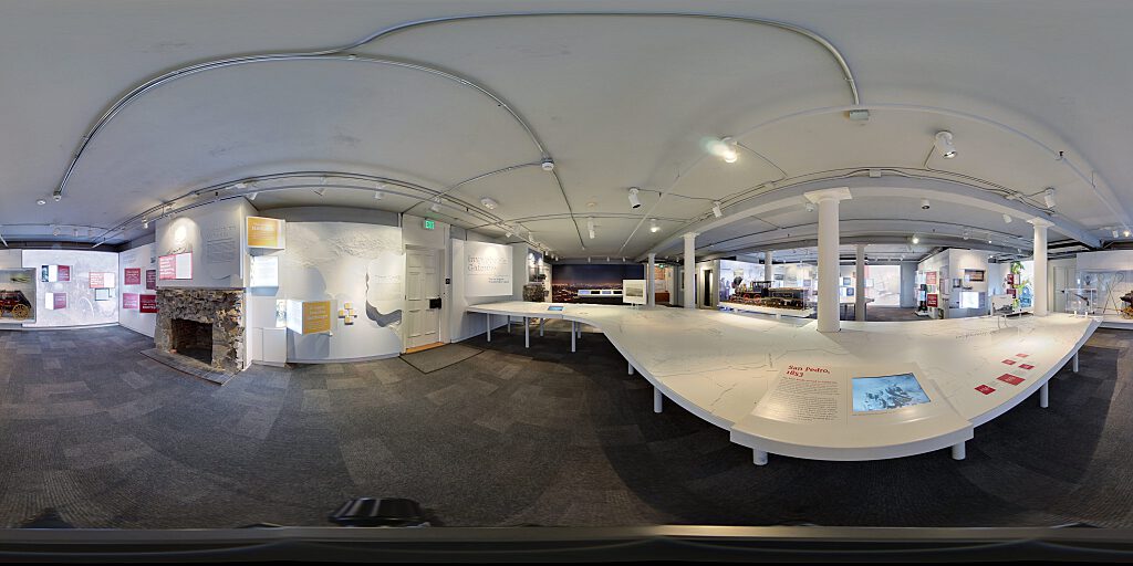 Panoramic view of Improbable Gateway Exhibit showing large wall photos, pop out text panels and in-depth descriptions of the transportation story of the Los Angeles Harbor. Artifacts include a tall ship replica and a model steam train.