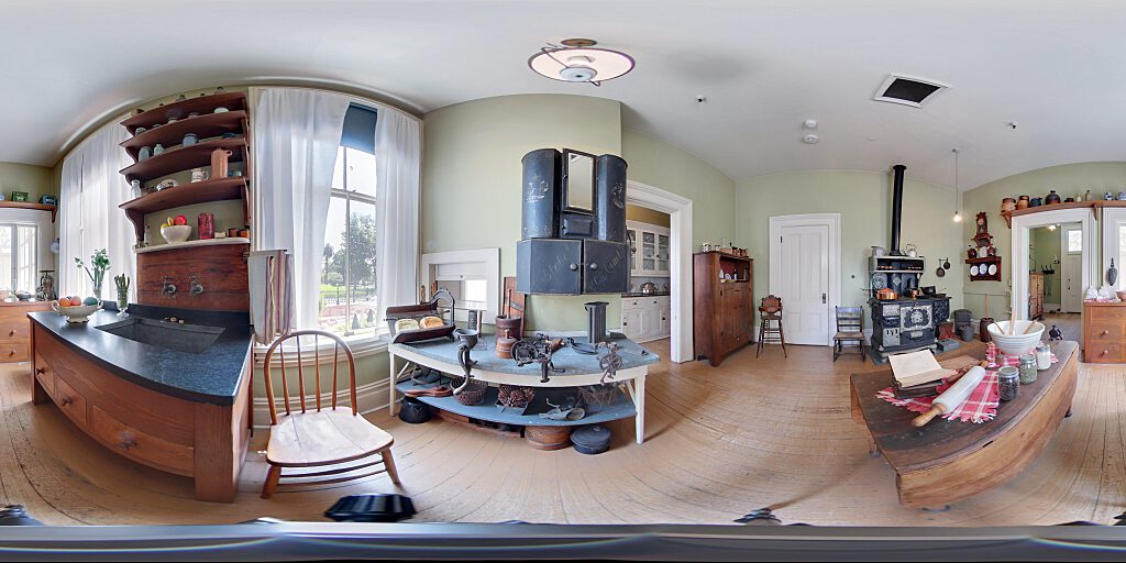 Panoramic view of the kitchen showing black stove, spice cabinets, Victorian Era gadgets and wooden table in center of room