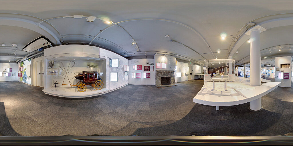 Panoramic view of Improbable Gateway Exhibit showing large wall photos, pop out text panels and in-depth descriptions of the transportation story of the Los Angeles Harbor. Artifacts include a tall ship replica and a model steam train and stage coach.