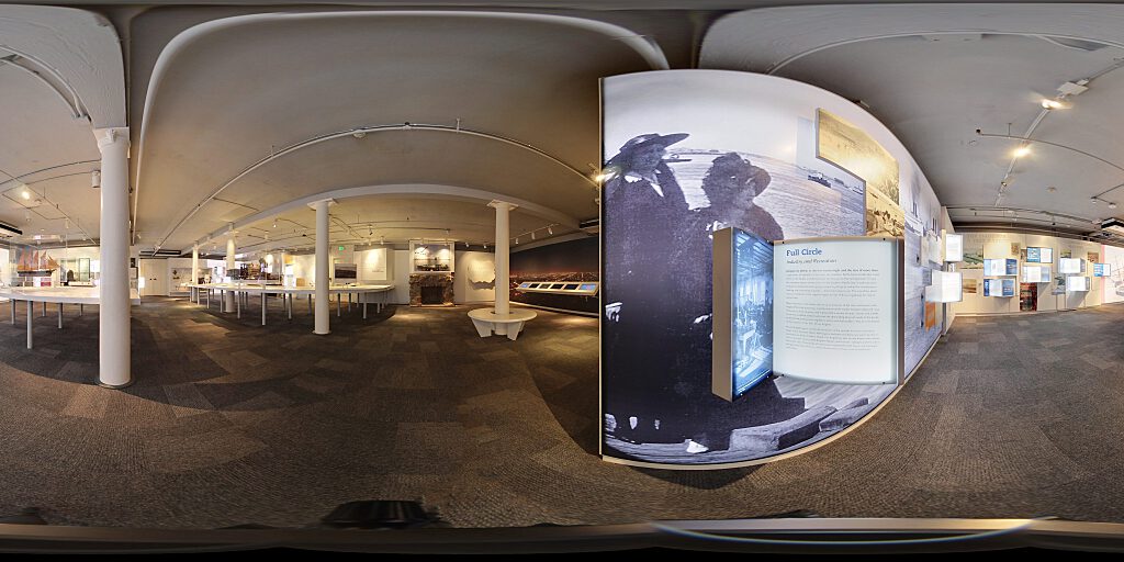 Panoramic view of Improbable Gateway Exhibit showing large wall photos, pop out text panels and in-depth descriptions of the transportation story of the Los Angeles Harbor. Artifacts include a tall ship replica and a model steam train.