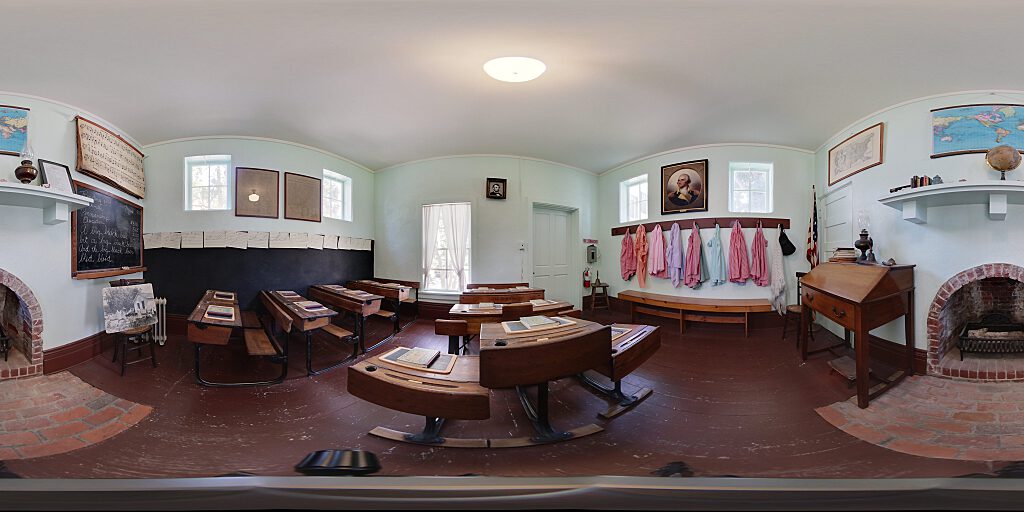 Panoramic view of Schoolhouse interior with desks with books, pinafores, and fireplace.