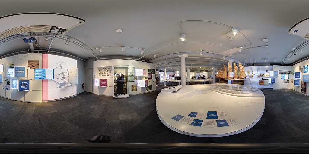 Panoramic view of Improbable Gateway Exhibit showing large wall photos, pop out text panels and in-depth descriptions of the transportation story of the Los Angeles Harbor. Artifacts include a tall ship replica and a model steam train.