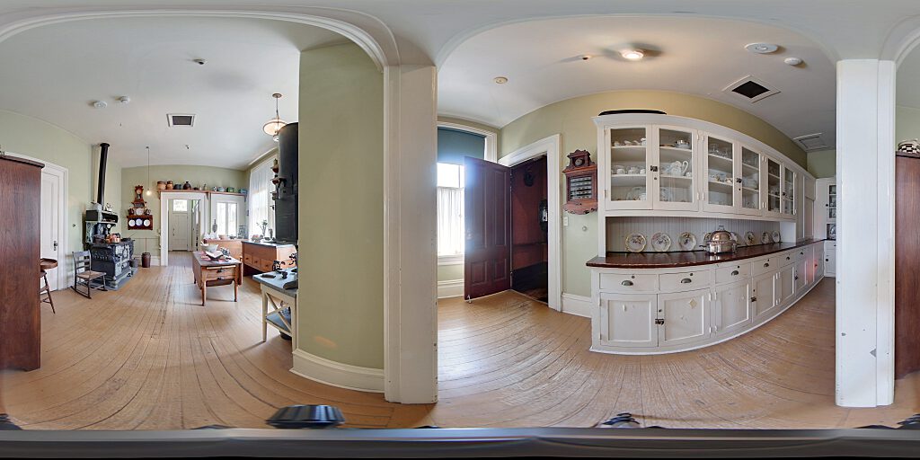 Panoramic view of the entrance to the kitchen showing Victorian Era call system next to white storage cabinets with long view of black stove