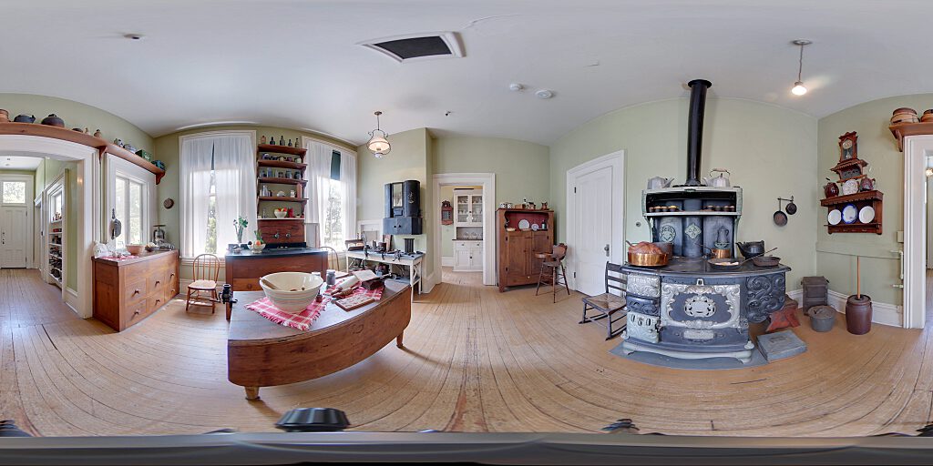 Panoramic view of the kitchen showing black stove, spice cabinets, Victorian Era gadgets and wooden table in center of room