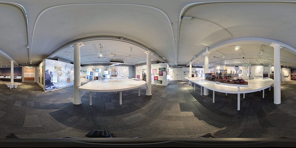 Panoramic view of Improbable Gateway Exhibit showing large wall photos, pop out text panels and in-depth descriptions of the transportation story of the Los Angeles Harbor. Artifacts include a tall ship replica and a model steam train.