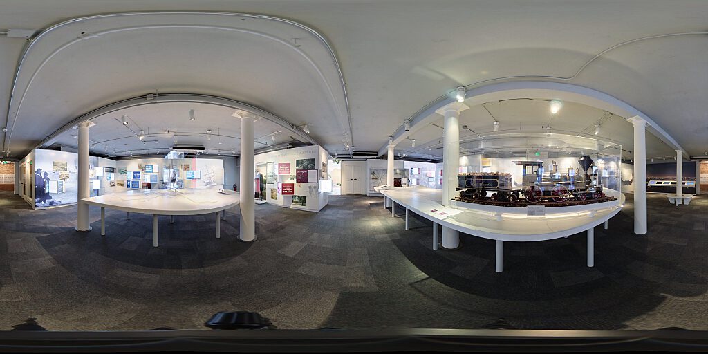 Panoramic view of Improbable Gateway Exhibit showing large wall photos, pop out text panels and in-depth descriptions of the transportation story of the Los Angeles Harbor. Artifacts include a tall ship replica and a model steam train.