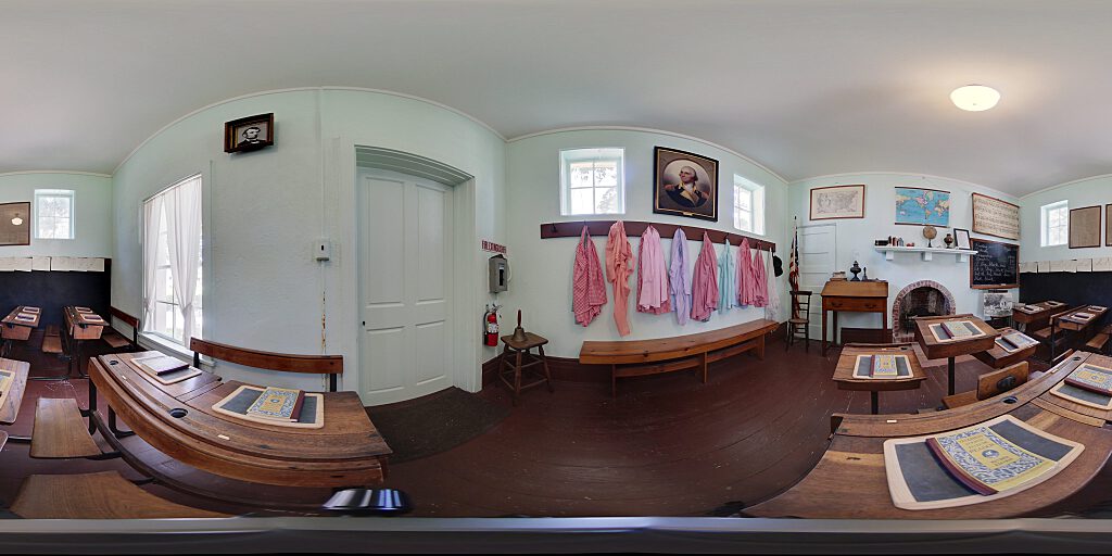 Panoramic view of Schoolhouse interior with desks with books, pinafores, and fireplace.